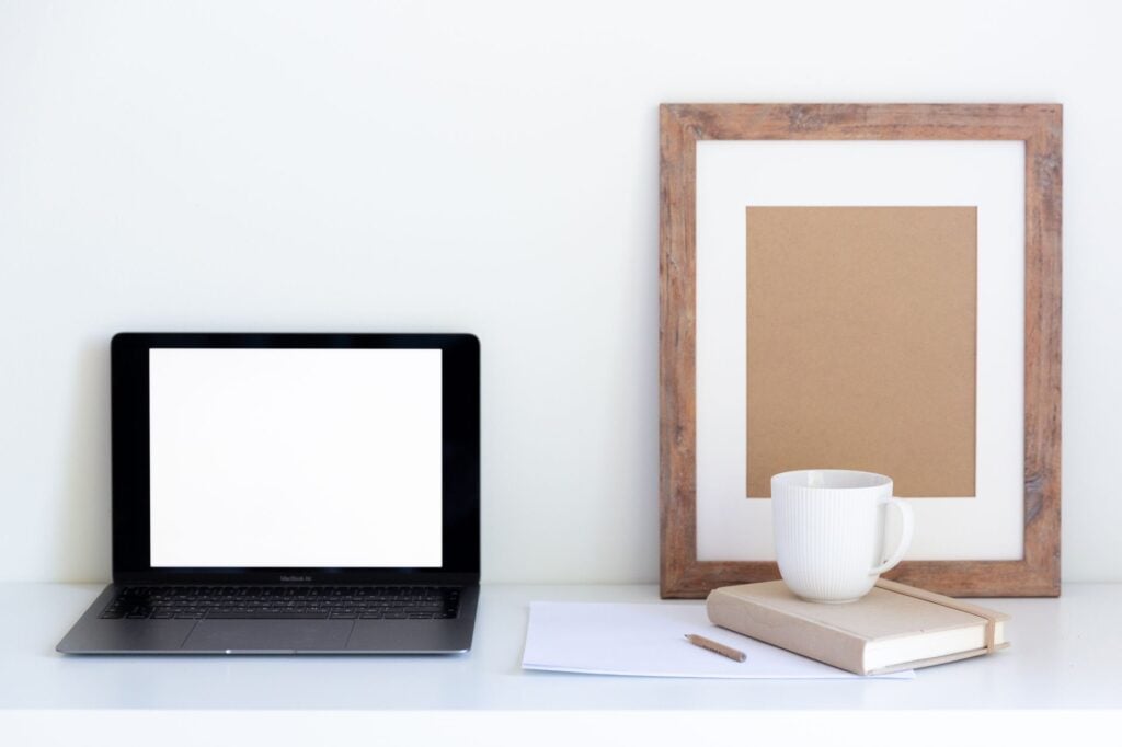 Empty frame placed near cup of coffee on closed notepad and white paper with empty screen of computer aside against white background