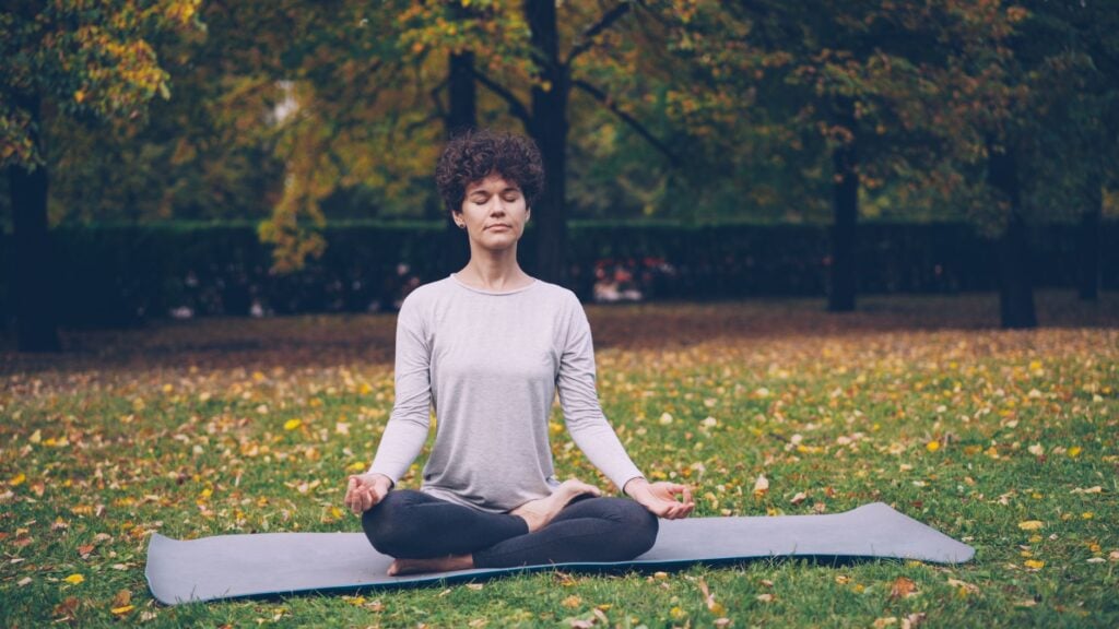 A woman practices meditation on a mat in a tranquil autumn park setting.