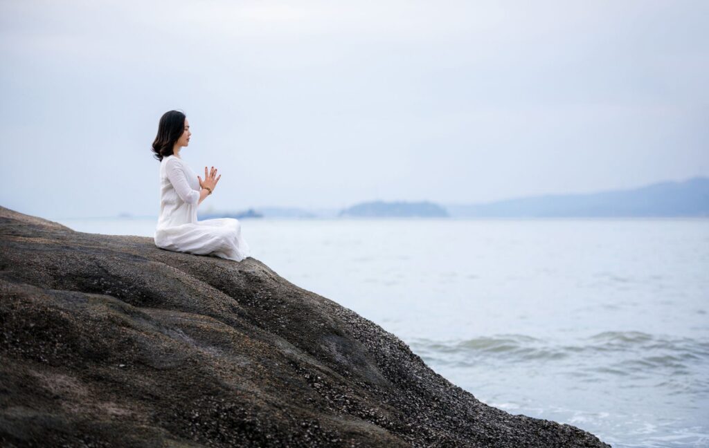 A woman meditating in a white dress by the serene sea, embodying peace and wellness.
