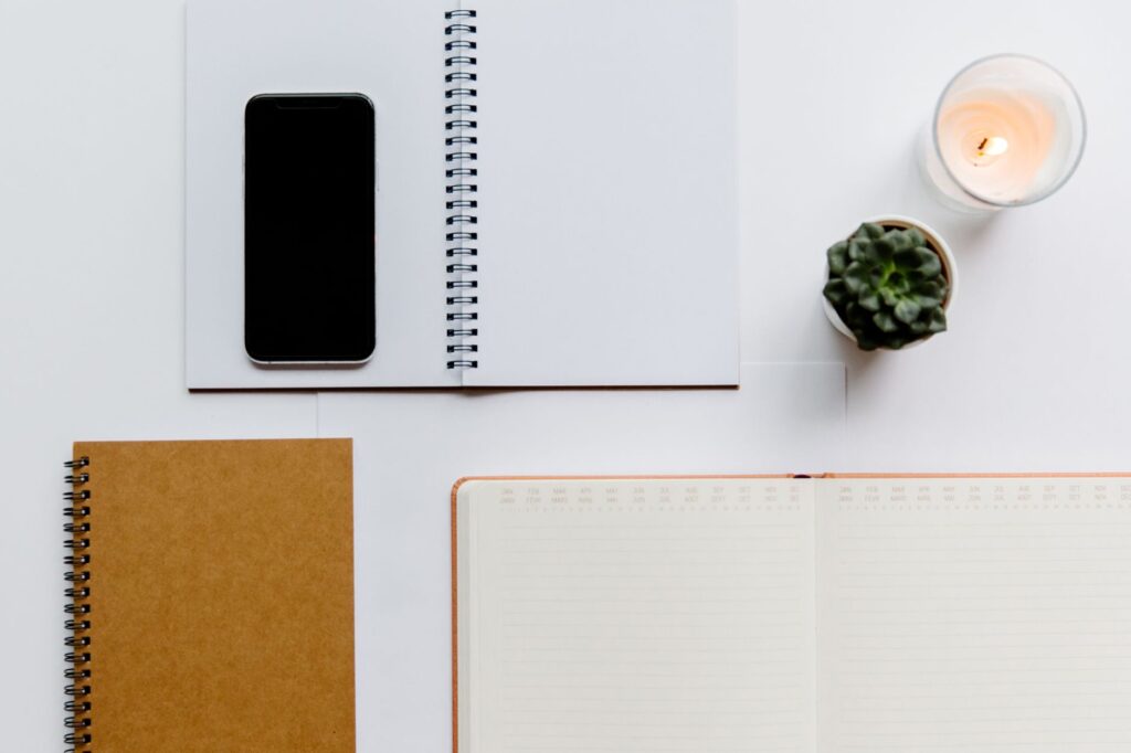 Aerial view of a minimalist office setup featuring notebooks, a smartphone, candle, and succulent on a white background.