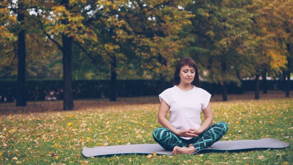How Can I Manage Anxiety-Related Physical Tension and Muscle Pain? 2 A woman practicing meditation on a yoga mat in a serene autumn park setting.