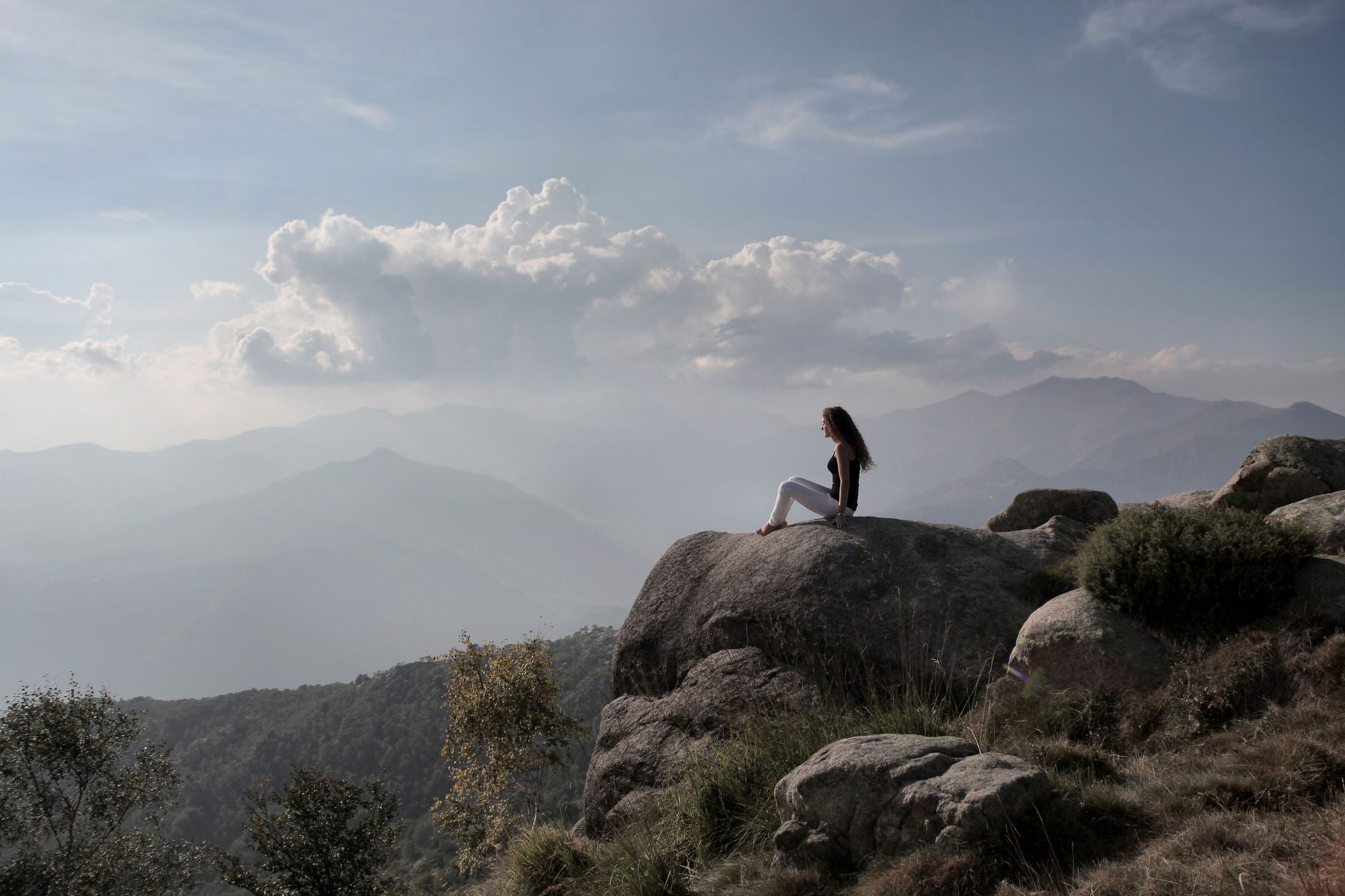 Blog 4 A woman seated on a rock enjoys a panoramic view of a mountainous landscape under a cloudy sky.