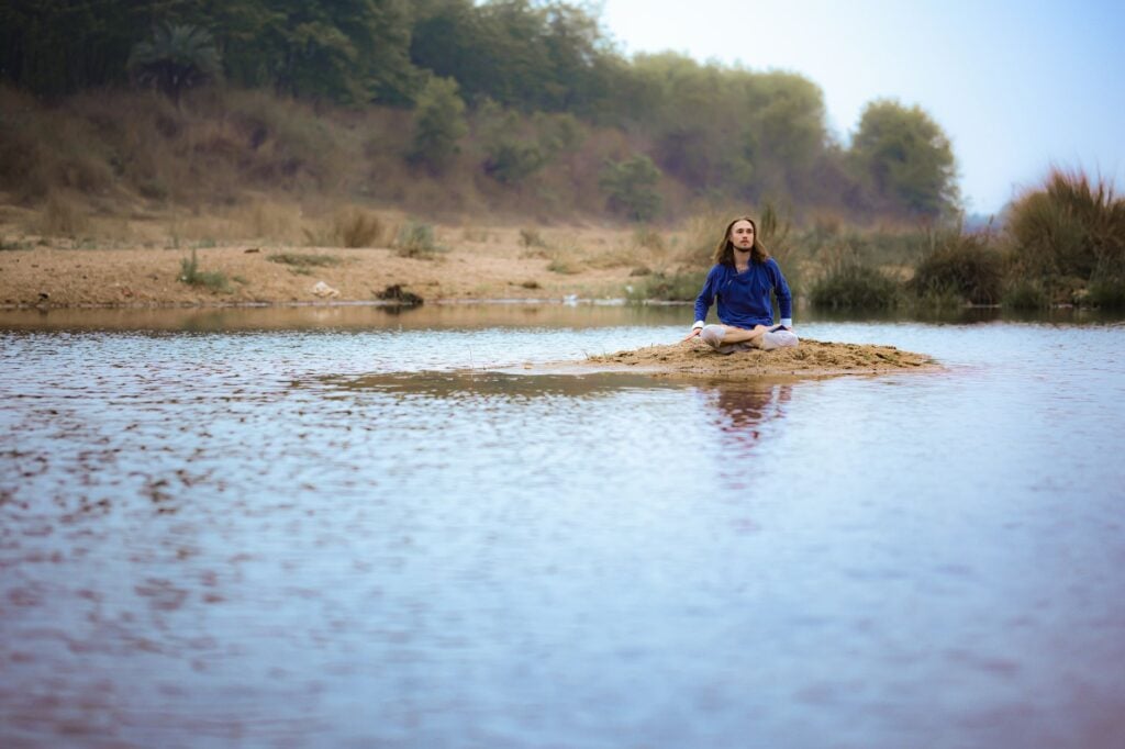 Person meditating on a sandy island in a serene river, surrounded by lush greenery in Dumka, India.