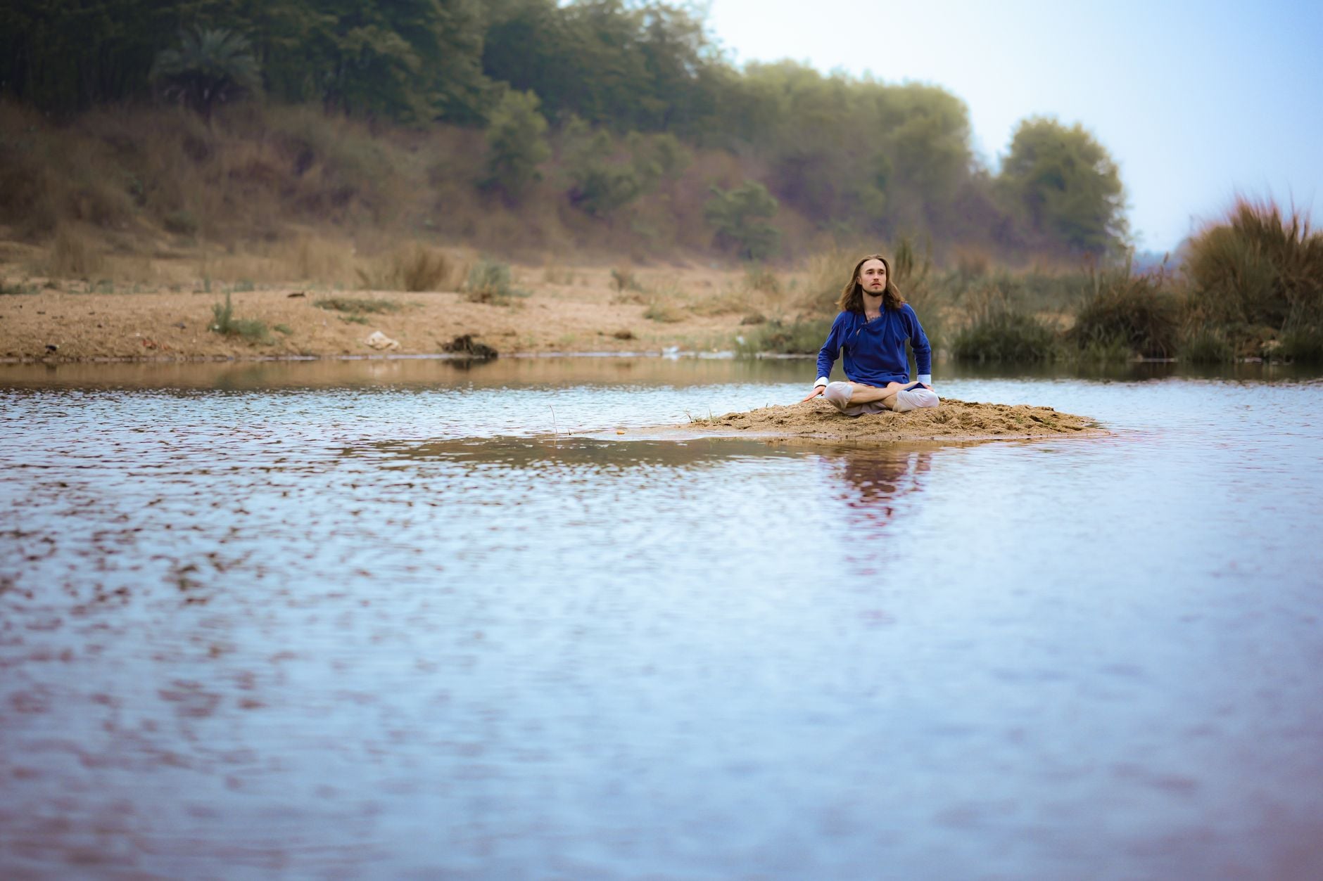 Blog 1 Person meditating on a sandy island in a serene river, surrounded by lush greenery in Dumka, India.