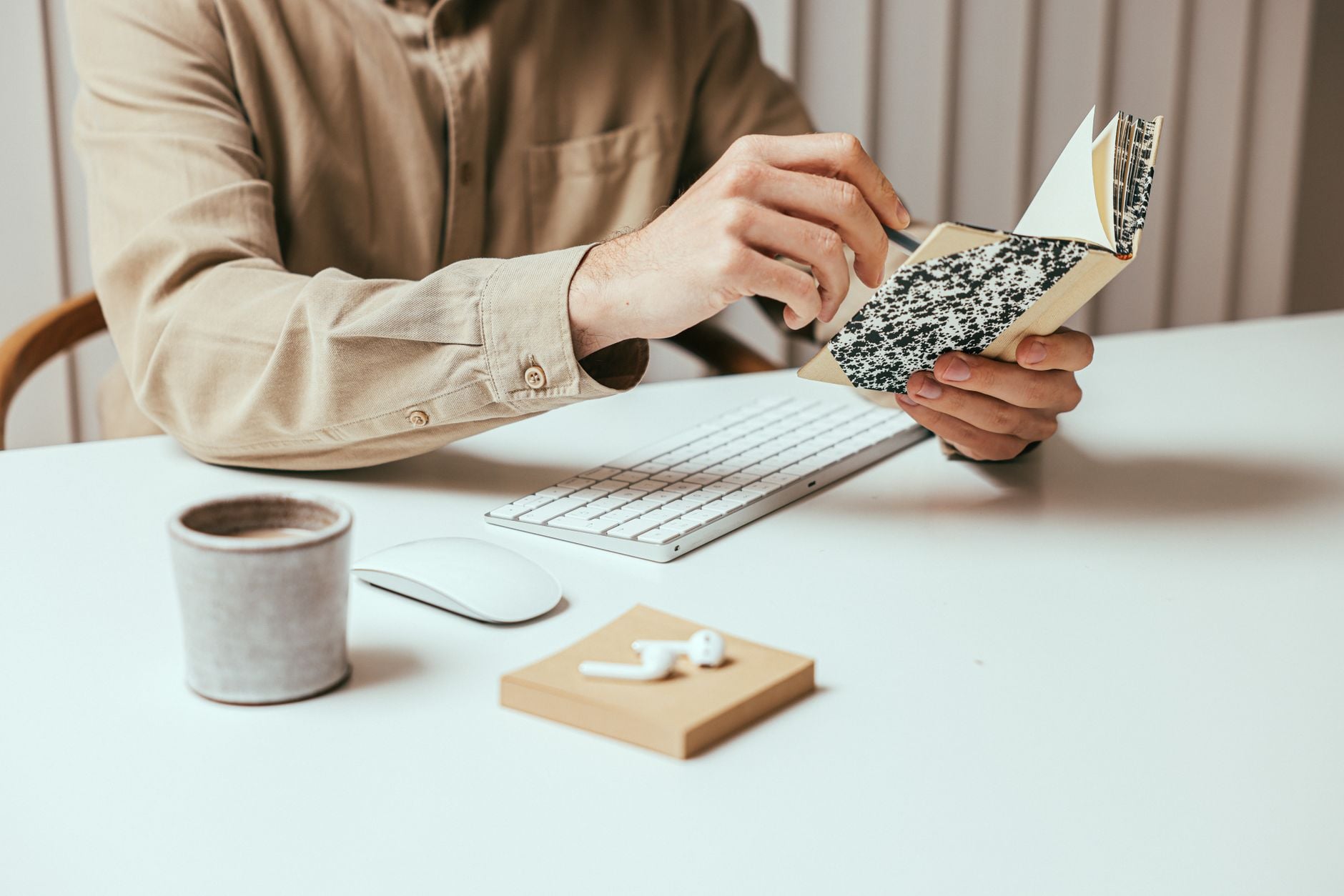 Close-up of a person holding a notepad at a modern desk with coffee, keyboard, and earbuds.