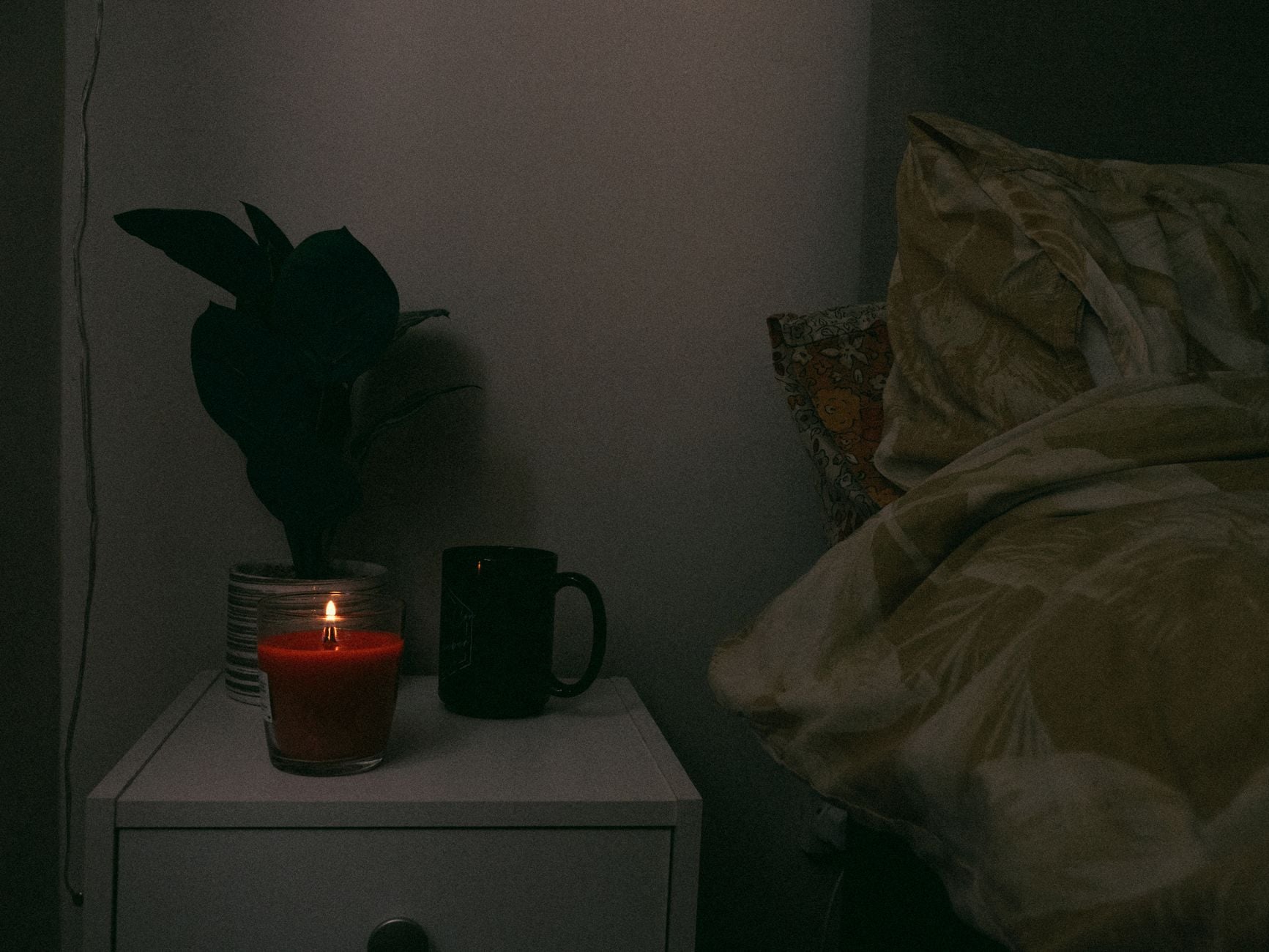 A cozy nightstand scene with a candle, mug, and plant beside a bed, creating a warm atmosphere.