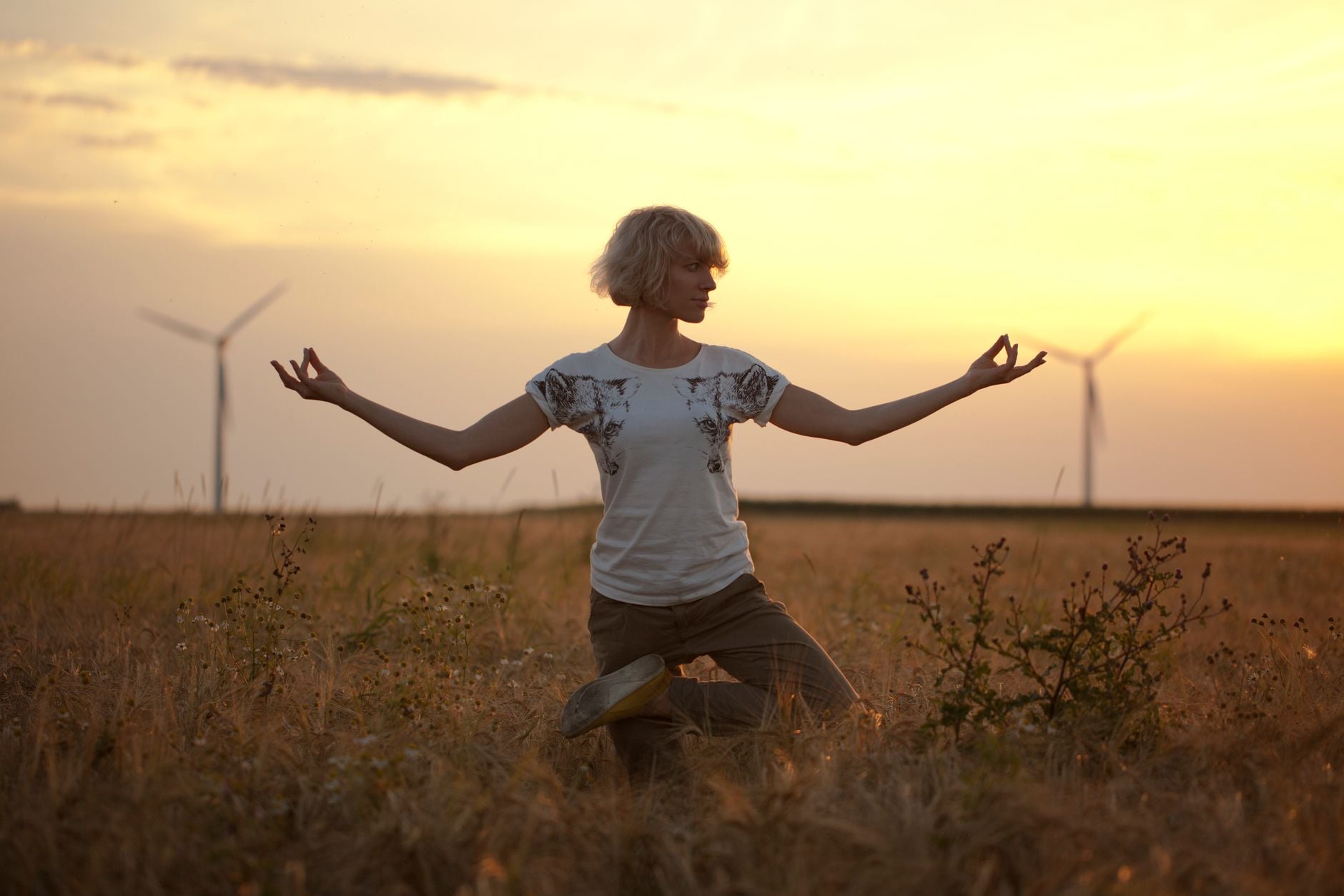 A woman practices yoga in a grassy field during sunset with wind turbines in the background.